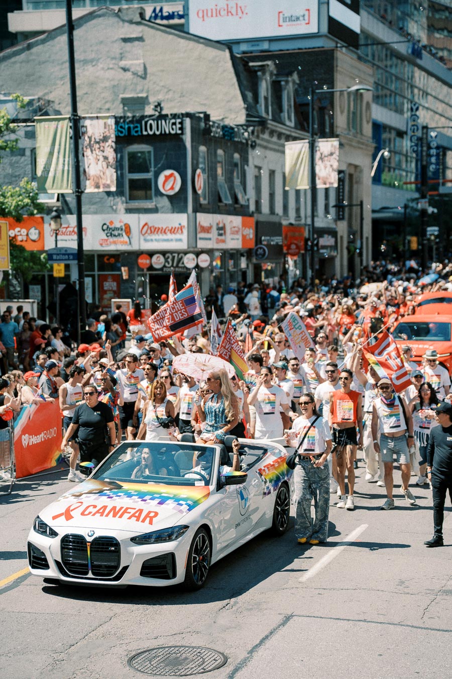 A lively parade featuring a white BMW convertible adorned with rainbow colors and the CANFAR logo, surrounded by a diverse crowd celebrating on a city street.