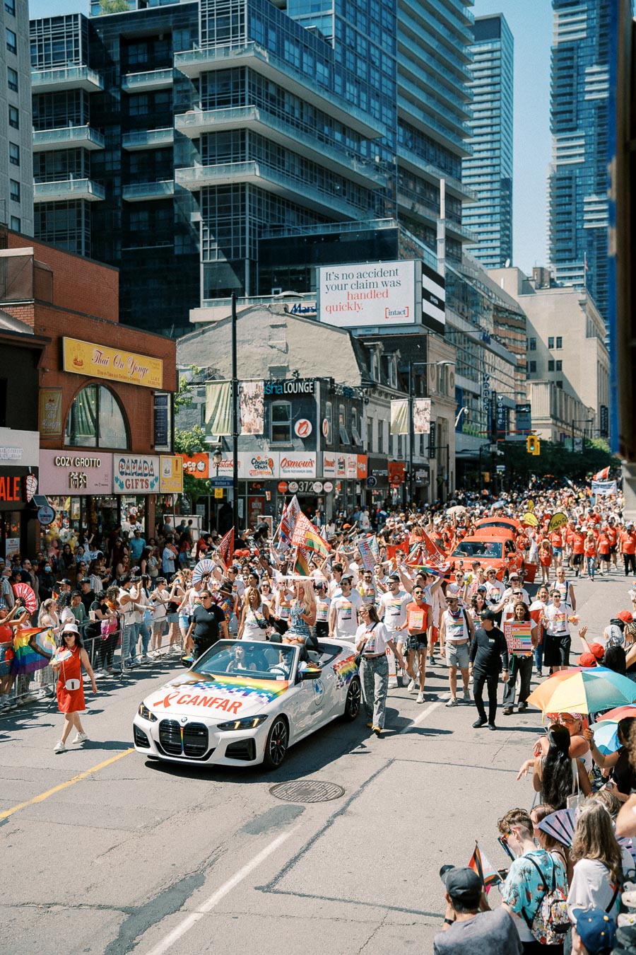 A lively parade featuring a white convertible with rainbow decorations and 'CANFAR' written on it, surrounded by a cheering crowd on a bustling city street.