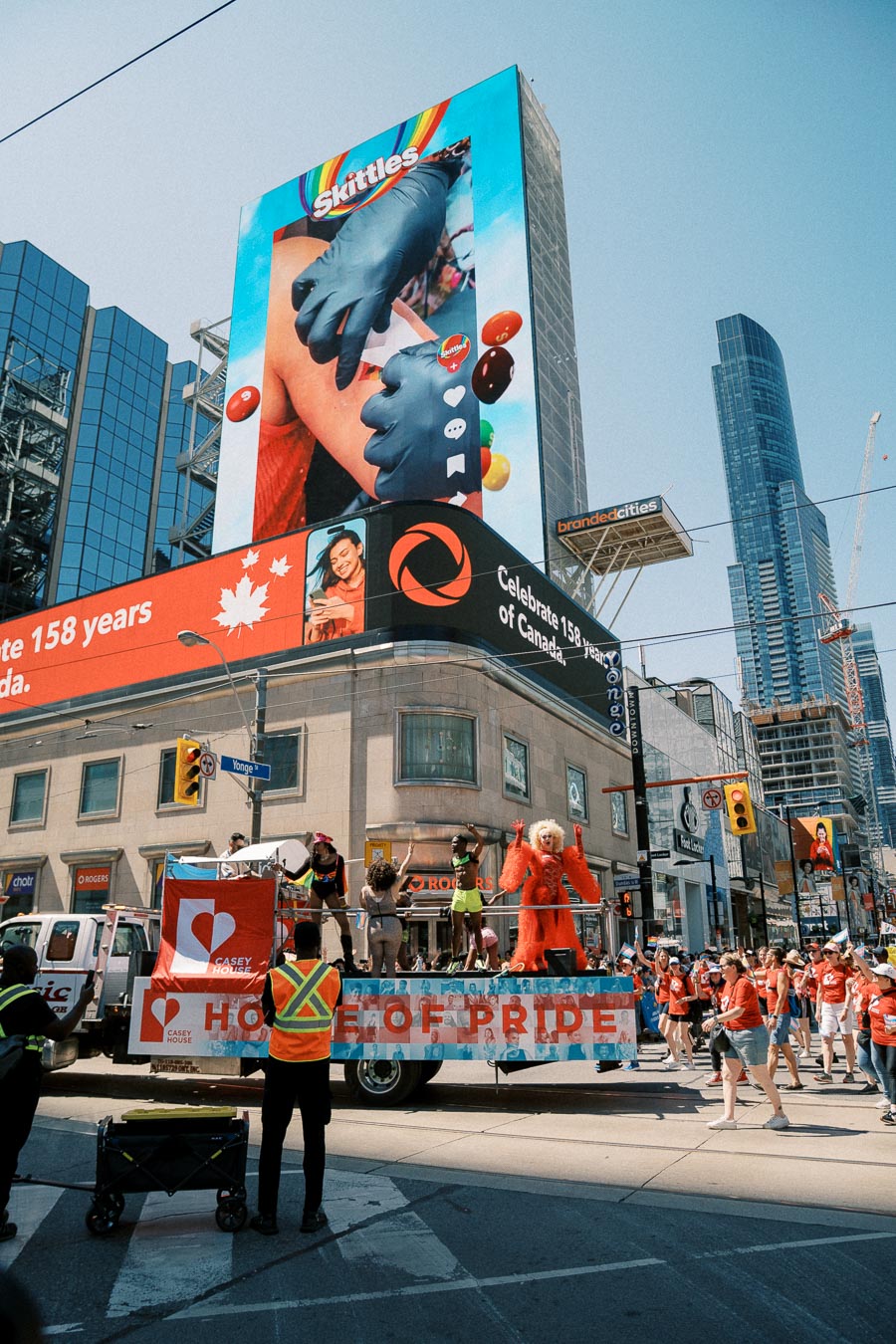A vibrant parade scene in a city with a float labeled 'House of Pride' and large digital billboards, including a Skittles advertisement, in the background.