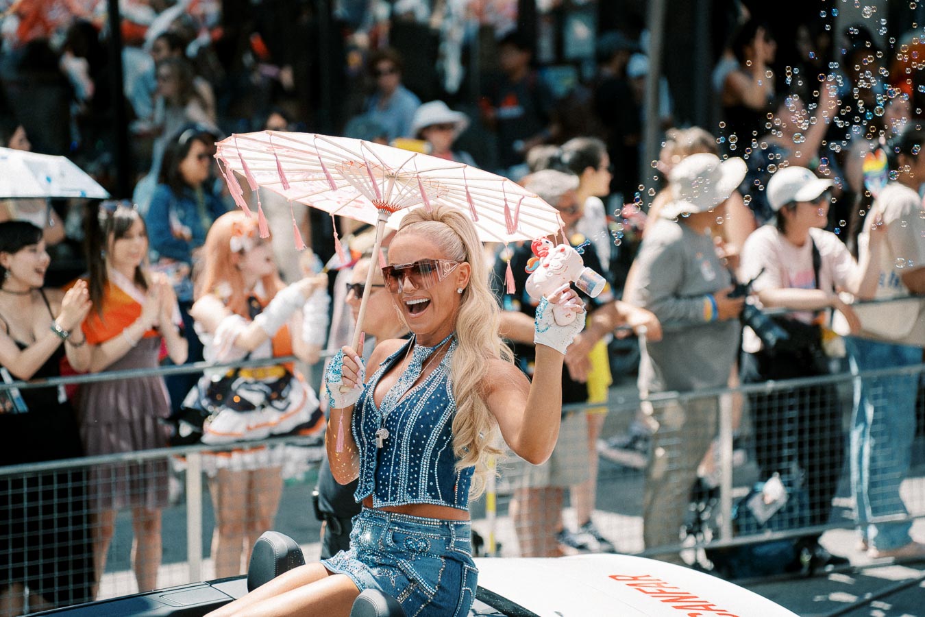 A vibrant scene at a parade featuring a person in a sparkling outfit holding a parasol and a bubble gun, surrounded by an enthusiastic crowd.