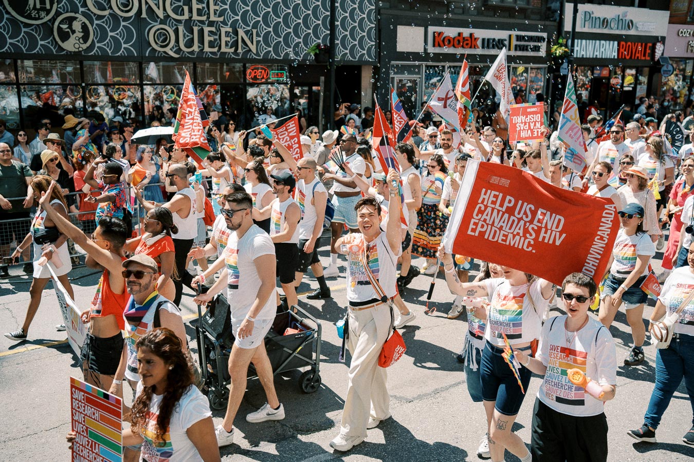 Participants in a pride parade march with signs promoting HIV awareness and research in Canada.