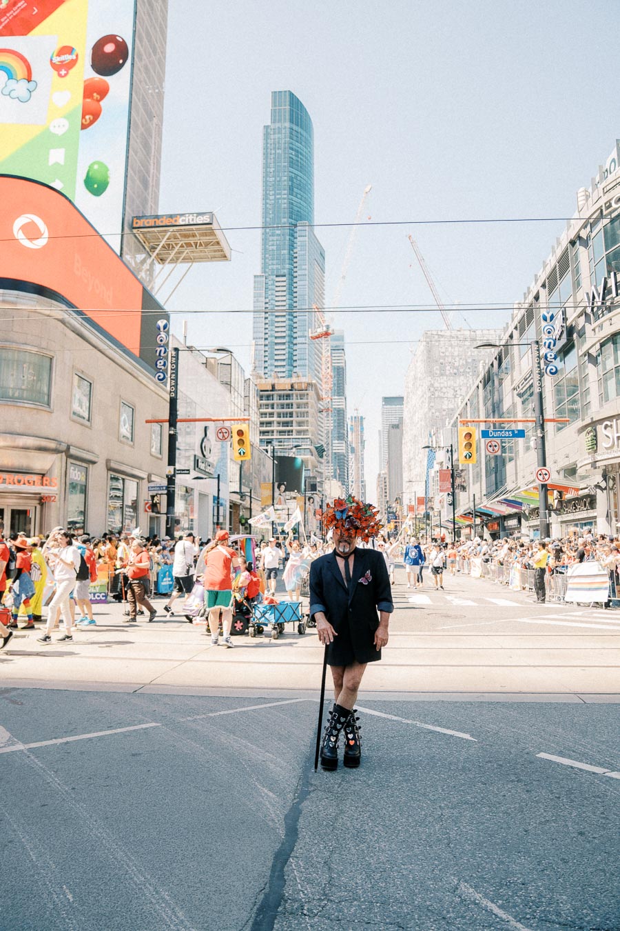 A vibrant parade scene in a bustling city, featuring a person in a unique outfit with a colorful headpiece, surrounded by crowds and skyscrapers.