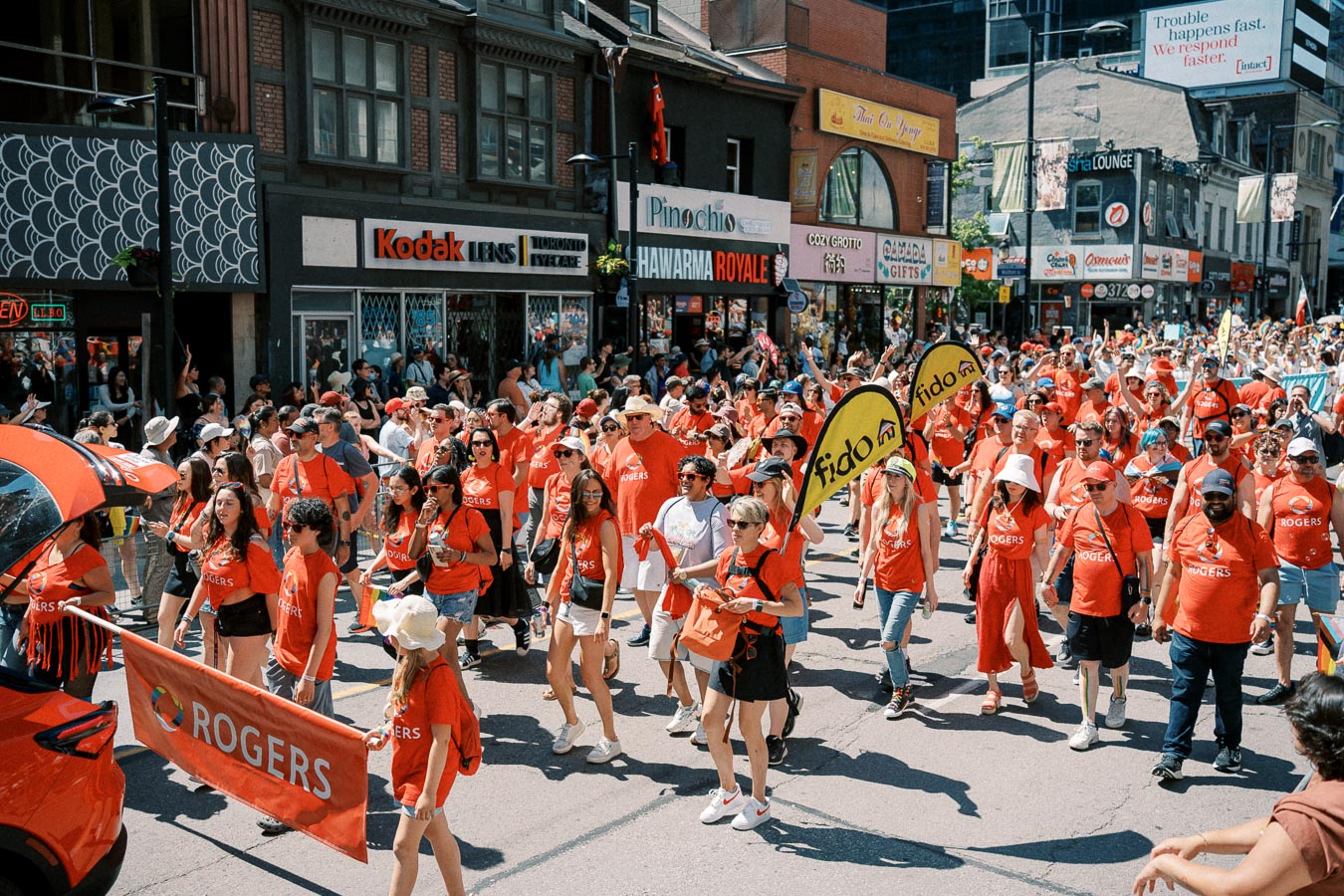 A vibrant parade featuring participants in red Rogers shirts marching down a bustling city street, surrounded by onlookers and storefronts.