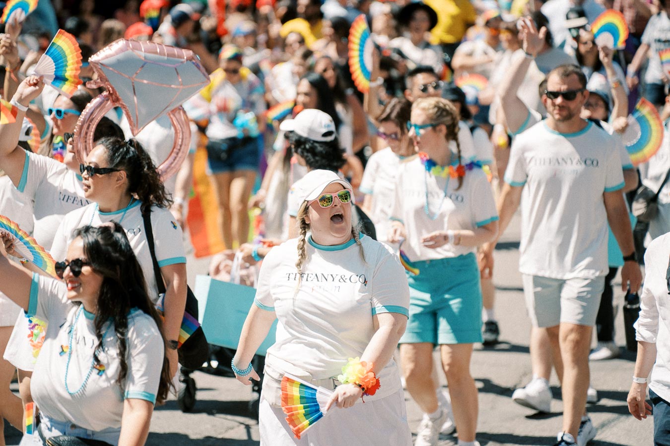 Participants in a parade wearing Tiffany &amp; Co. shirts, celebrating with rainbow fans and accessories.