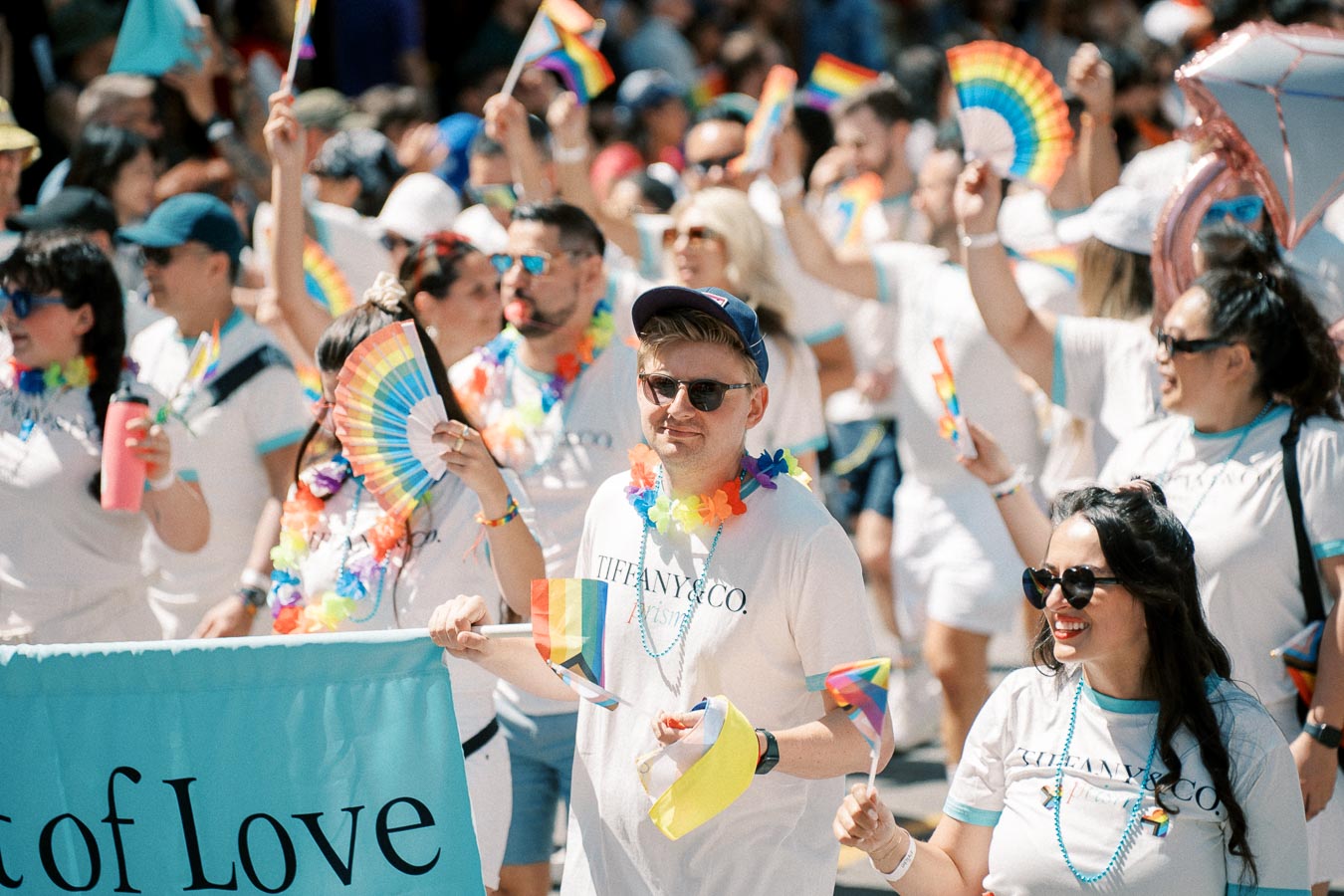 Participants in a pride parade wearing white shirts and rainbow accessories, celebrating with flags and fans.