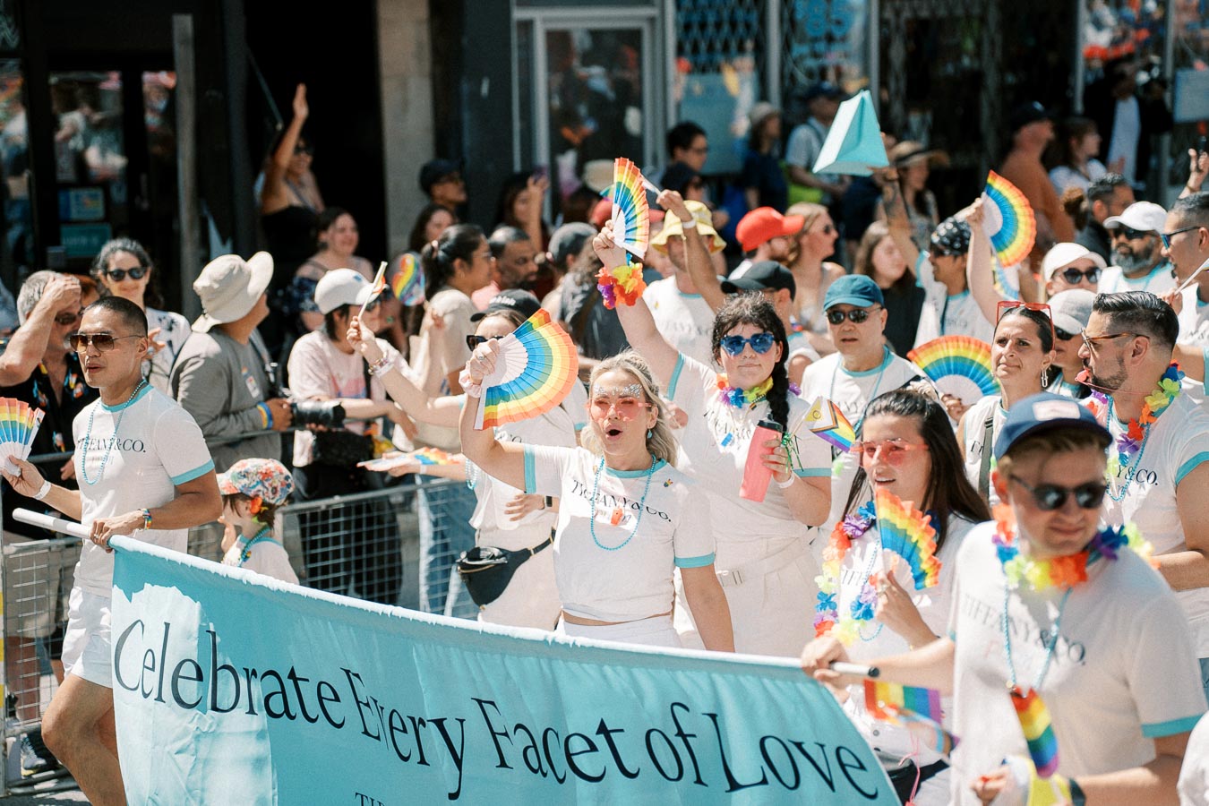 Participants in a pride parade celebrate with rainbow fans and colorful accessories, carrying a banner that reads 'Celebrate Every Facet of Love.'