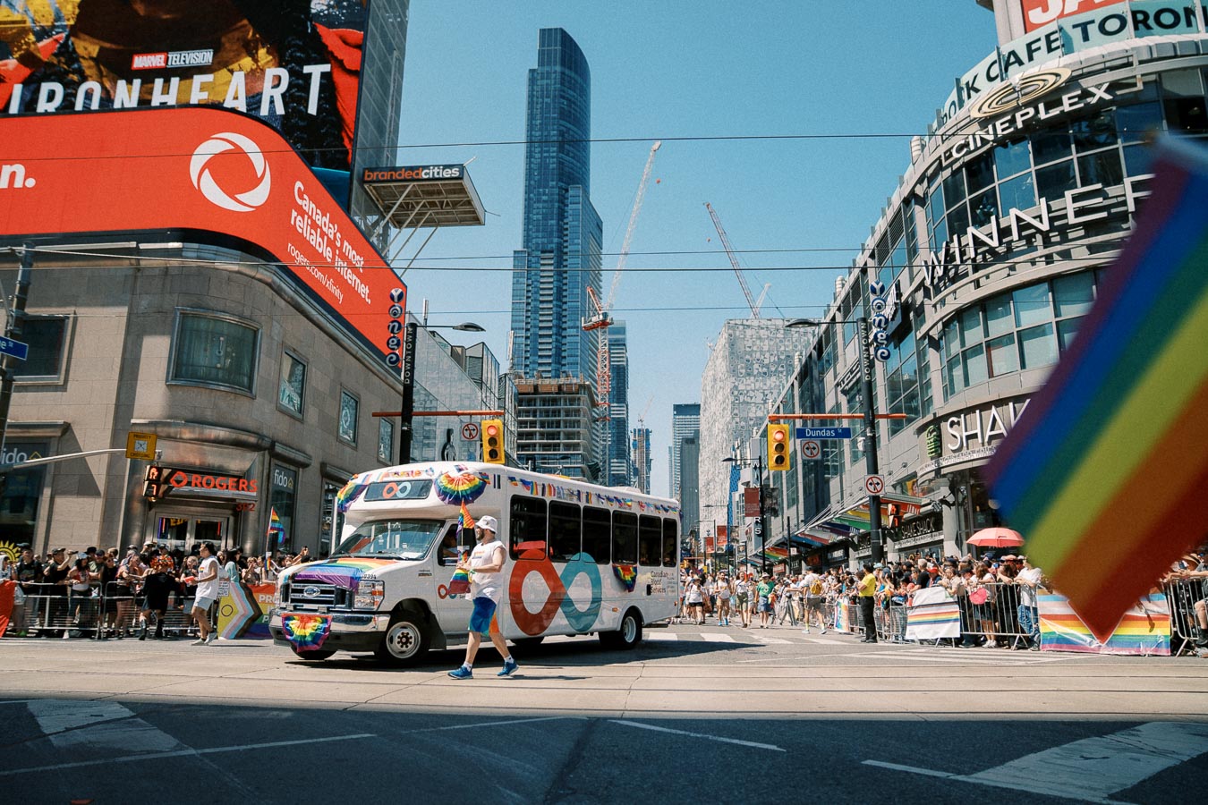 A pride parade in Toronto with a decorated bus and rainbow flags, set against the backdrop of city buildings and a large crowd.