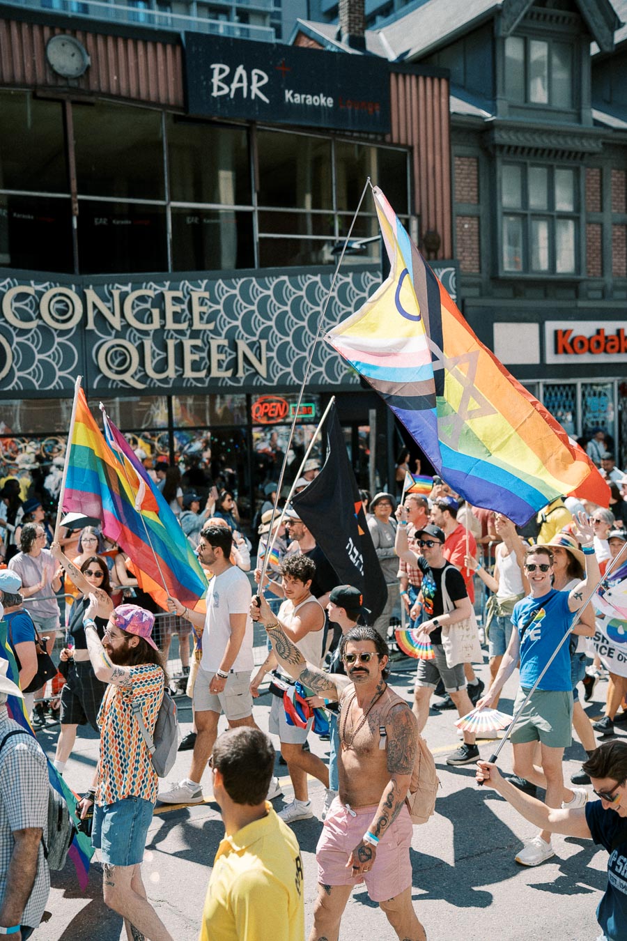 Participants in a pride parade march with rainbow flags in front of a building labeled 'Congee Queen'.