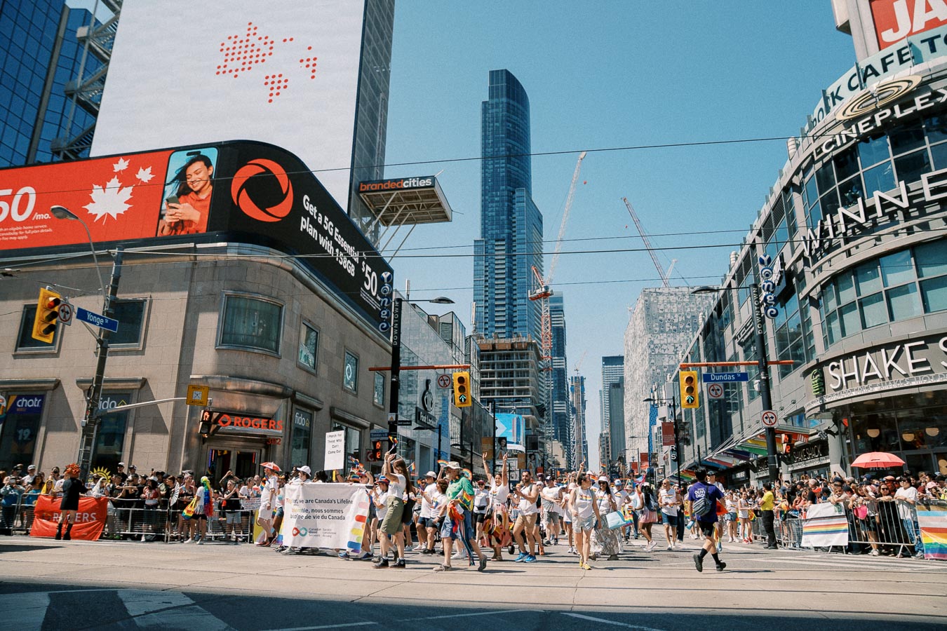 A vibrant parade takes place in downtown Toronto, featuring participants with banners and rainbow flags. The scene is set against a backdrop of tall buildings and digital billboards, capturing the lively urban atmosphere.