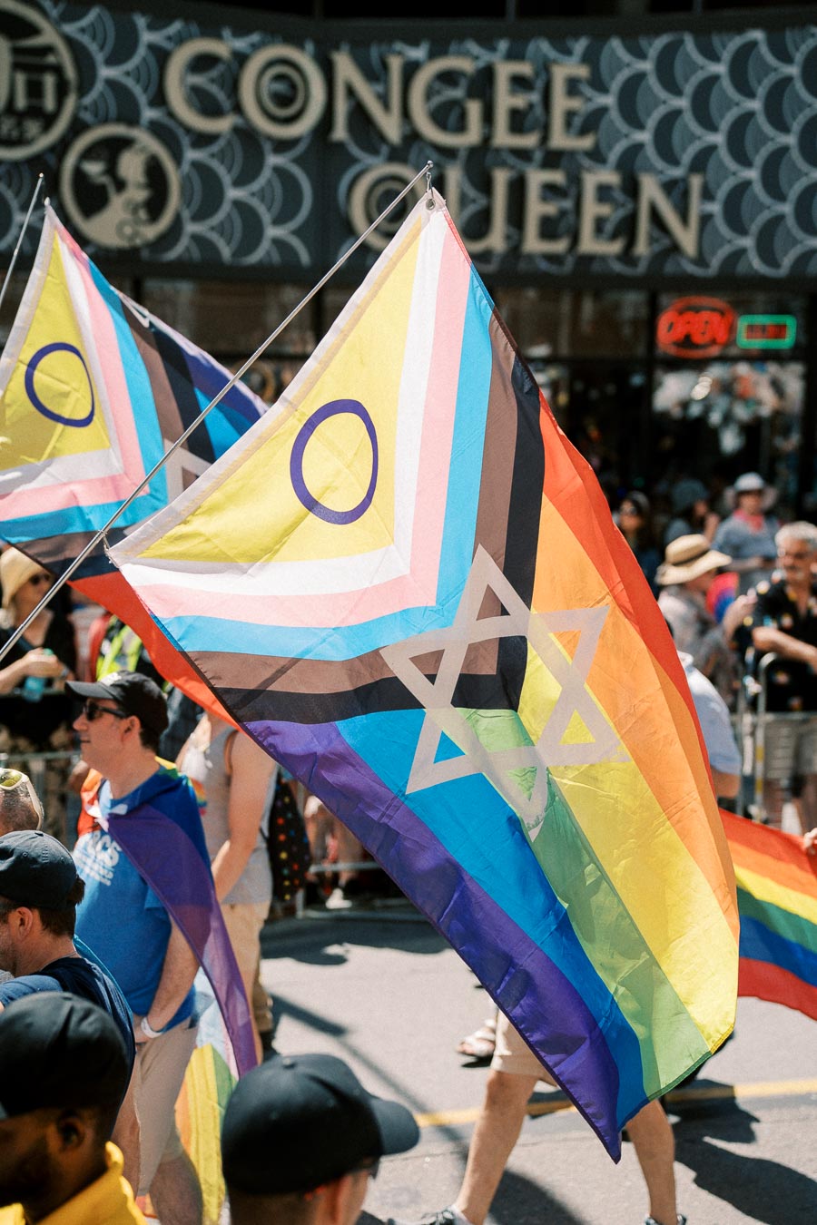 A vibrant pride flag featuring a Star of David and an intersex symbol is prominently displayed during a parade, with people and a restaurant sign in the background.