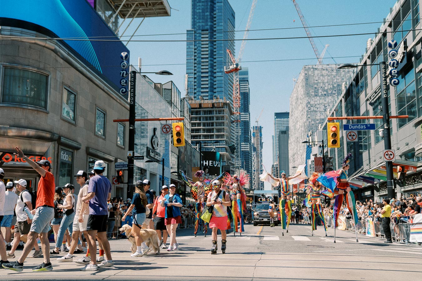 A lively parade takes place on a city street, featuring performers in colorful costumes on stilts. The event is attended by a large crowd, with skyscrapers and construction cranes visible in the background, indicating an urban setting.