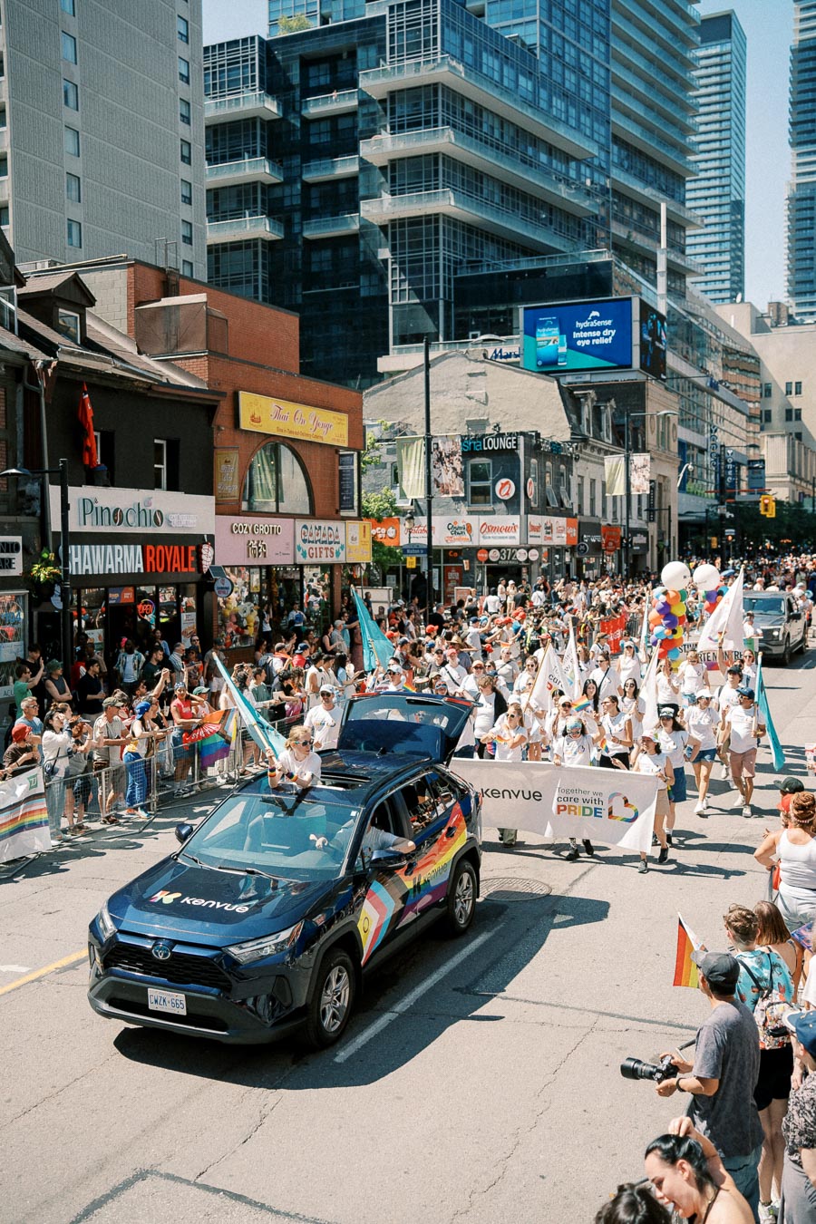 A lively pride parade takes place on a bustling city street, featuring a decorated car and participants holding colorful flags and banners, surrounded by cheering spectators.