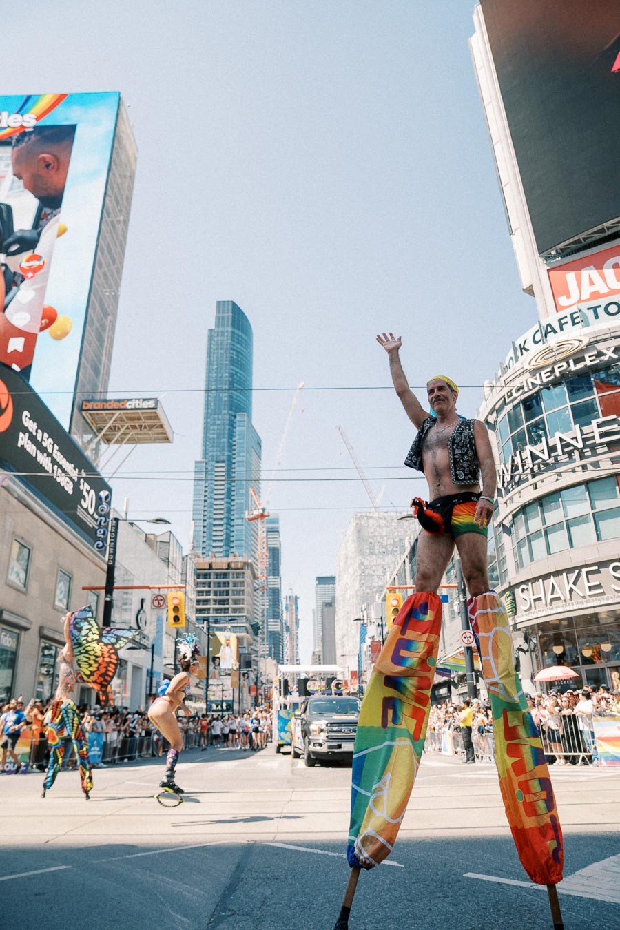 Performers on stilts entertain the crowd during a vibrant parade in a bustling city center, surrounded by skyscrapers and digital billboards.