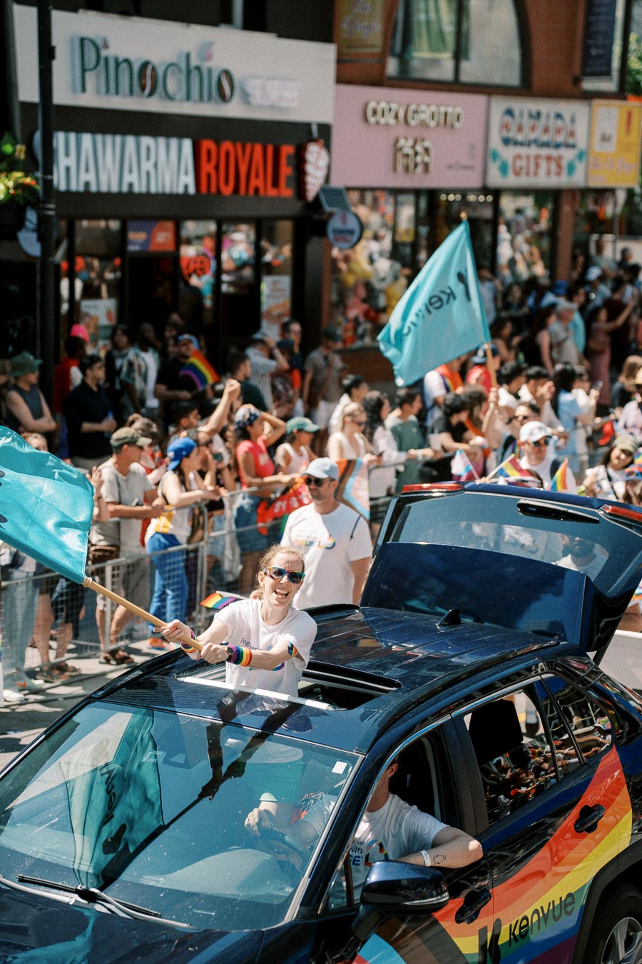 A lively parade scene with a person enthusiastically waving a teal flag from a car adorned with rainbow colors, surrounded by a cheering crowd and storefronts.