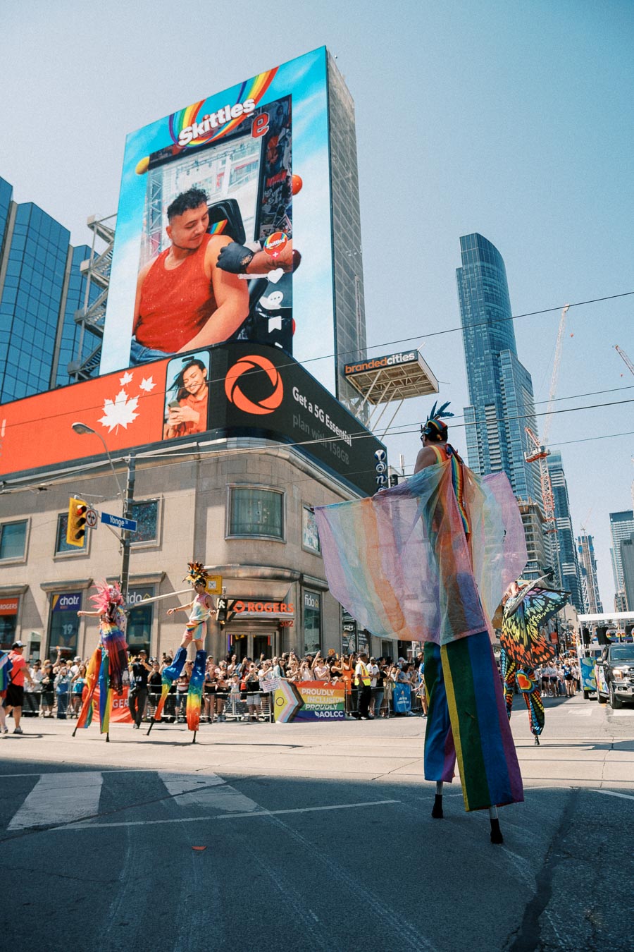 Performers on stilts in colorful costumes participate in a lively street parade, with a large Skittles advertisement displayed on a digital billboard in the background.