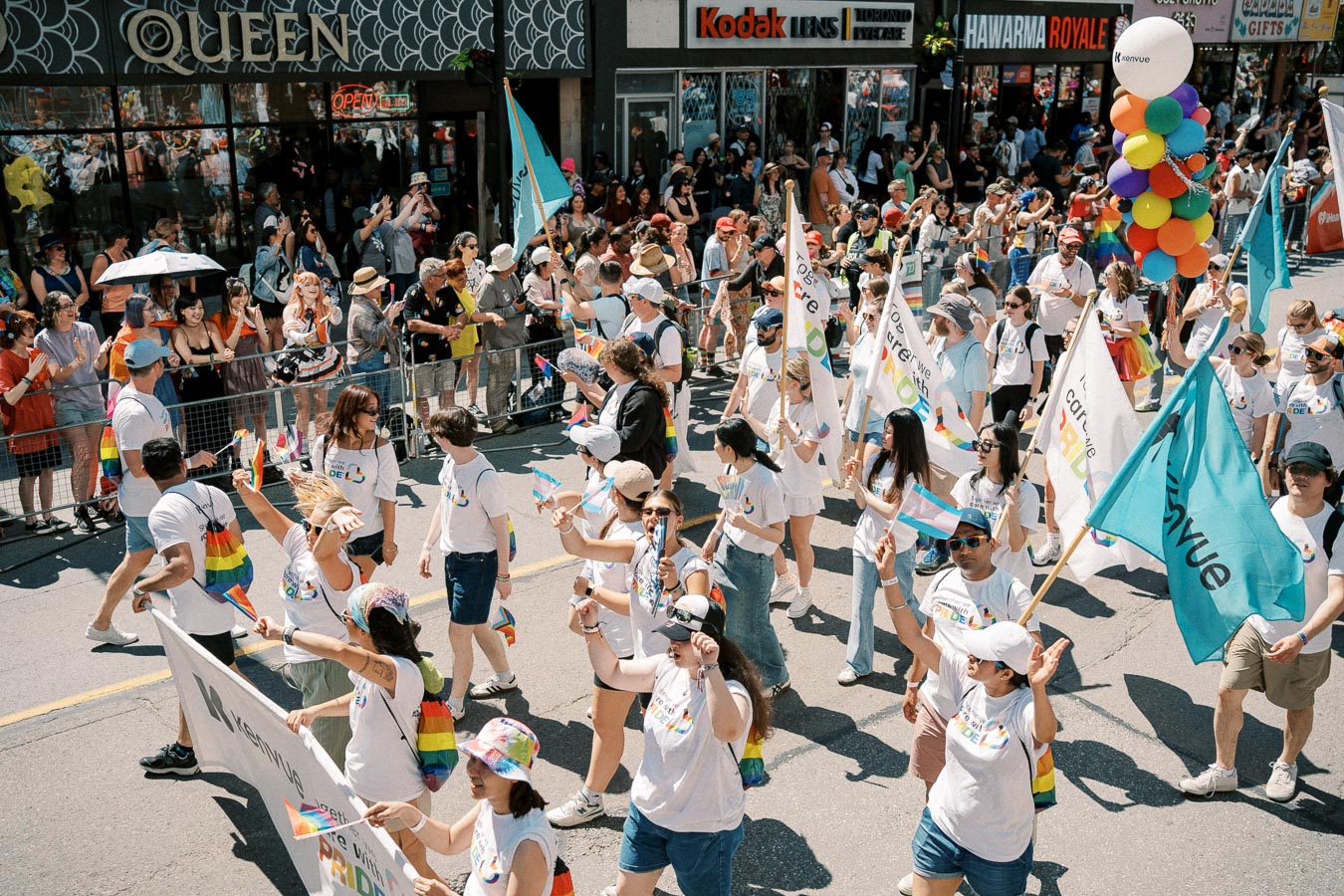 Participants in a pride parade march down a street, waving flags and carrying colorful balloons, while spectators cheer from the sidelines.