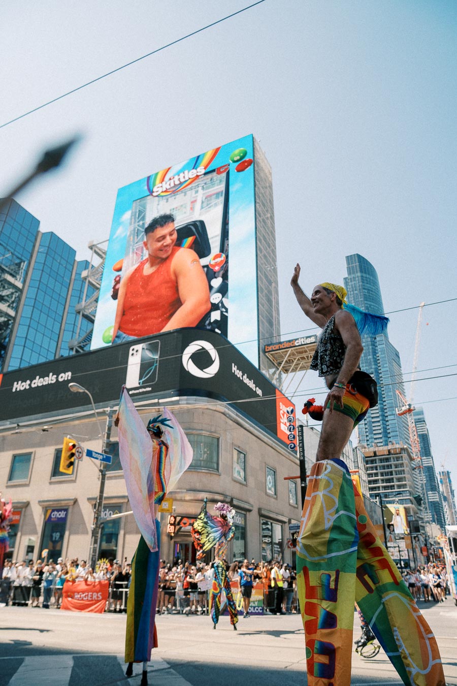 A lively parade scene featuring performers on stilts in colorful costumes, set against a backdrop of a large digital billboard in a bustling city.