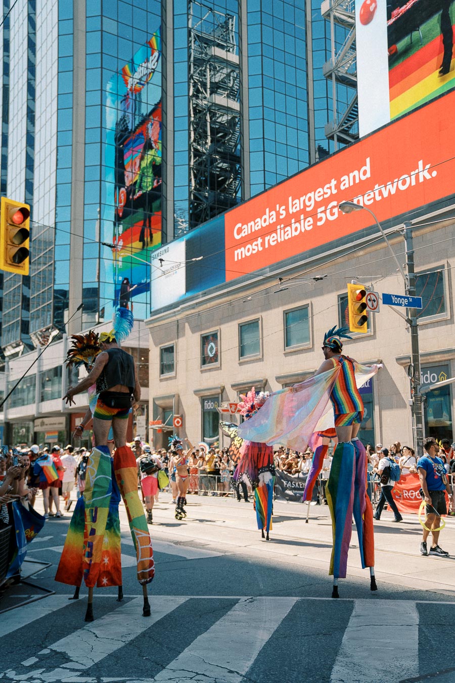 Participants in vibrant costumes on stilts parade through a city street during a festive event, with a large digital billboard in the background.