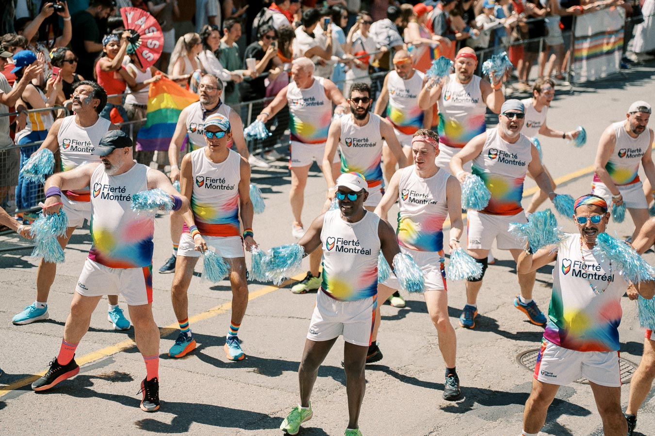 Participants in colorful rainbow outfits march with enthusiasm during the Fierté Montréal Pride Parade, holding blue pom-poms and celebrating diversity.