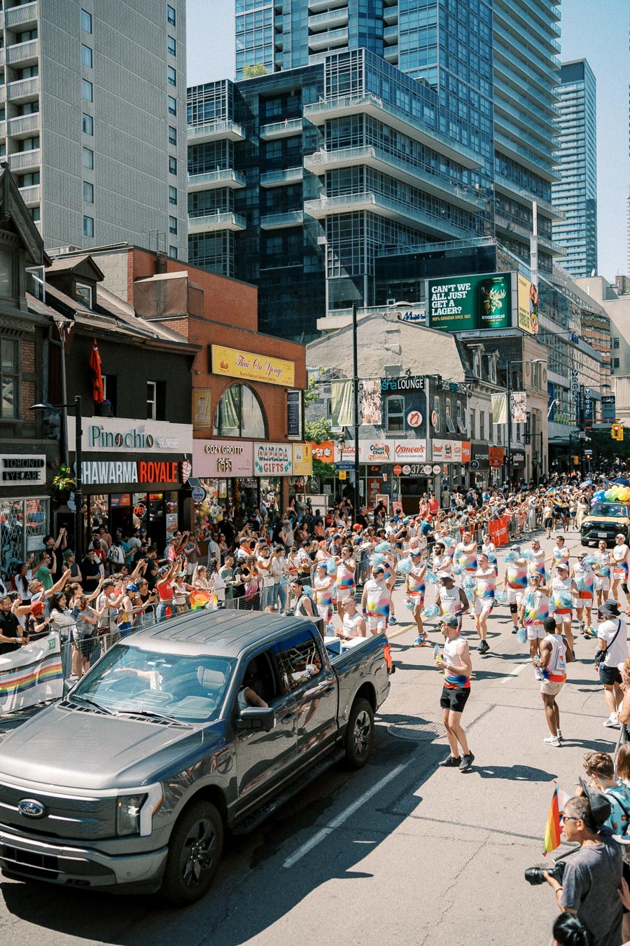 A vibrant street parade with participants in rainbow-themed outfits, watched by a cheering crowd. A Ford truck leads the procession, set against a backdrop of urban buildings.