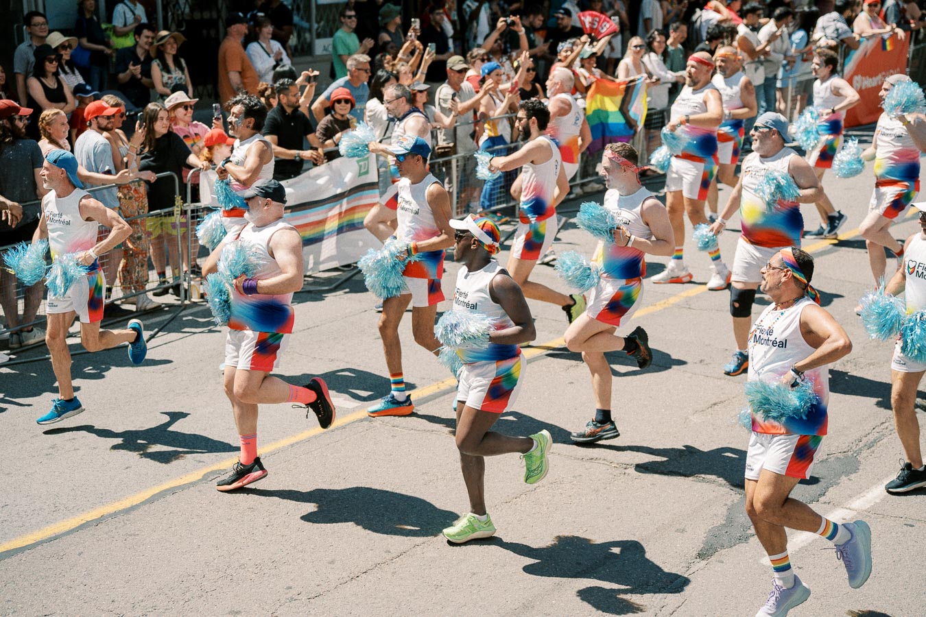 Participants in vibrant rainbow-themed outfits march enthusiastically in a pride parade, holding blue pom-poms, as spectators cheer them on from the sidelines.