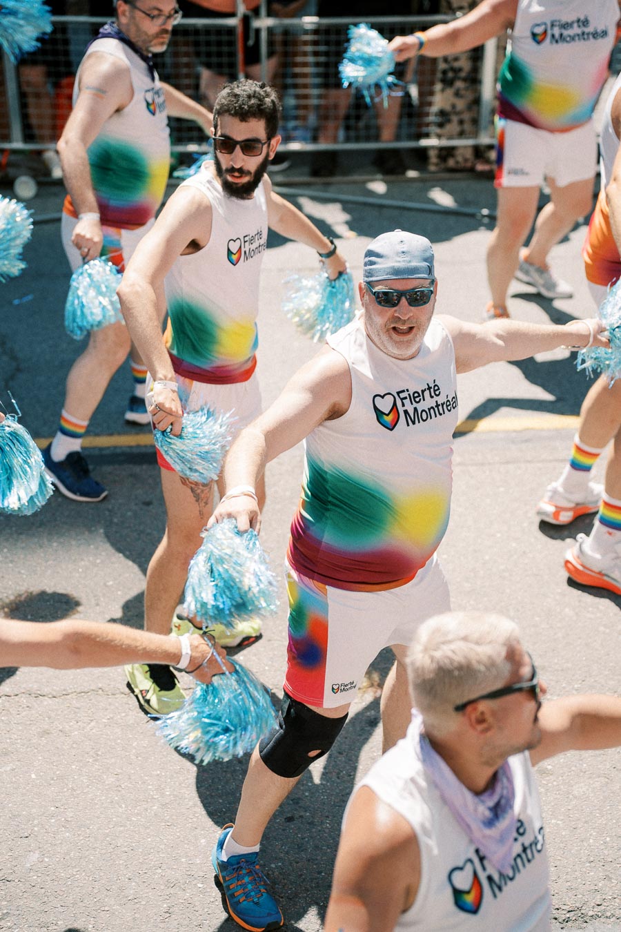 Participants in a pride parade wearing rainbow-themed outfits and holding blue pom-poms, celebrating diversity and inclusion.