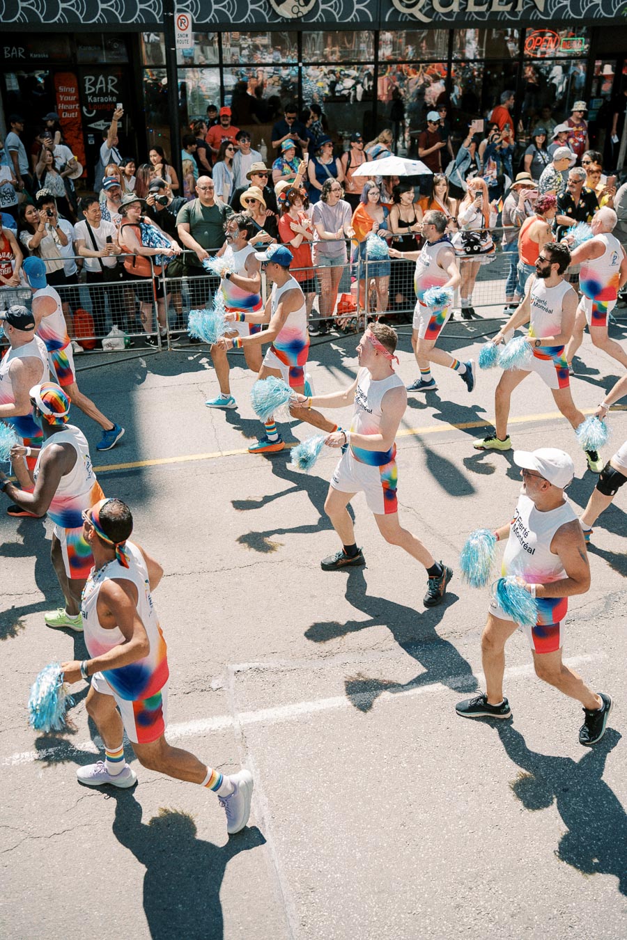 Participants in colorful outfits march in a parade, holding blue pom-poms, as spectators watch from the sidewalk.