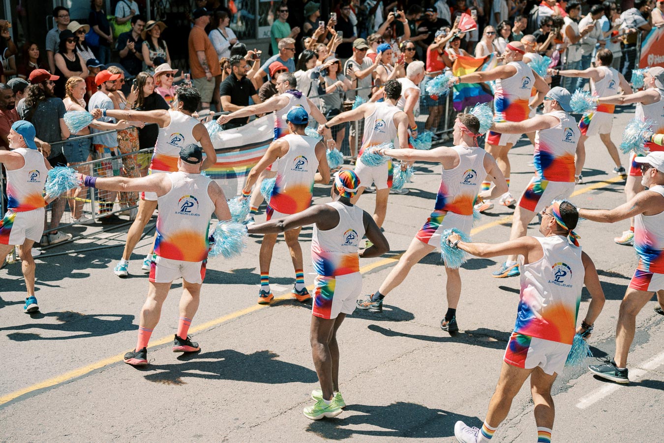 Participants in vibrant rainbow-themed outfits perform a synchronized routine during a lively parade, cheered on by enthusiastic spectators.