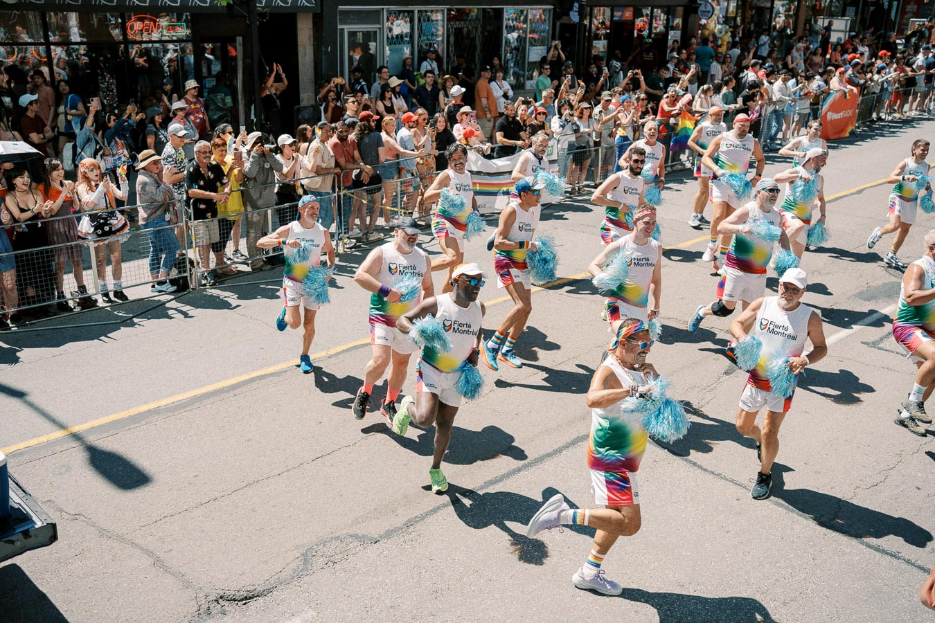Participants in vibrant rainbow outfits and blue pom-poms march energetically in a parade, cheered on by a lively crowd of spectators.