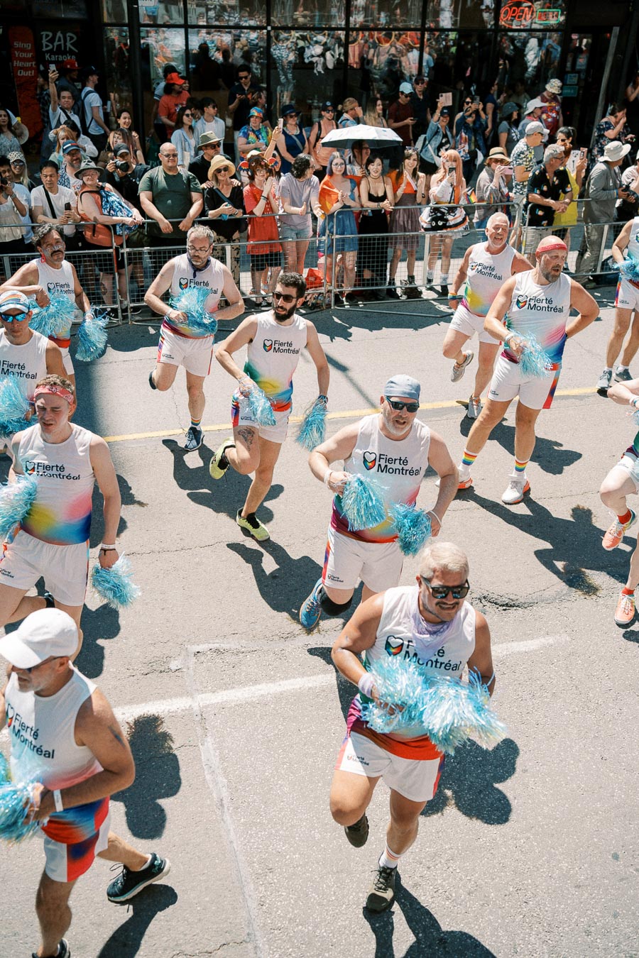 Participants in the Fierté Montréal parade energetically march down the street, waving blue pom-poms, as a lively crowd watches from the sidelines.