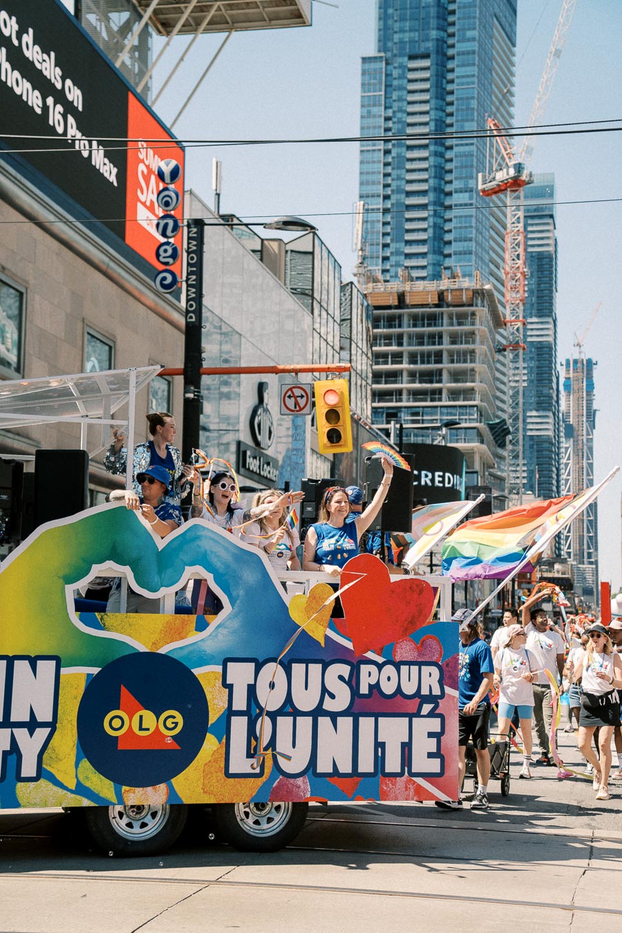 A vibrant parade float with participants celebrating and waving rainbow flags in a city street, surrounded by skyscrapers and construction cranes.