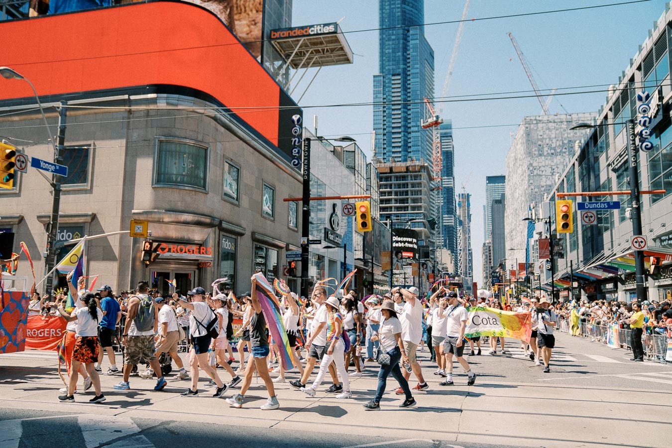 A lively parade takes place at the intersection of Yonge and Dundas Streets, featuring participants with rainbow flags and banners, set against a backdrop of urban skyscrapers and a cheering crowd.