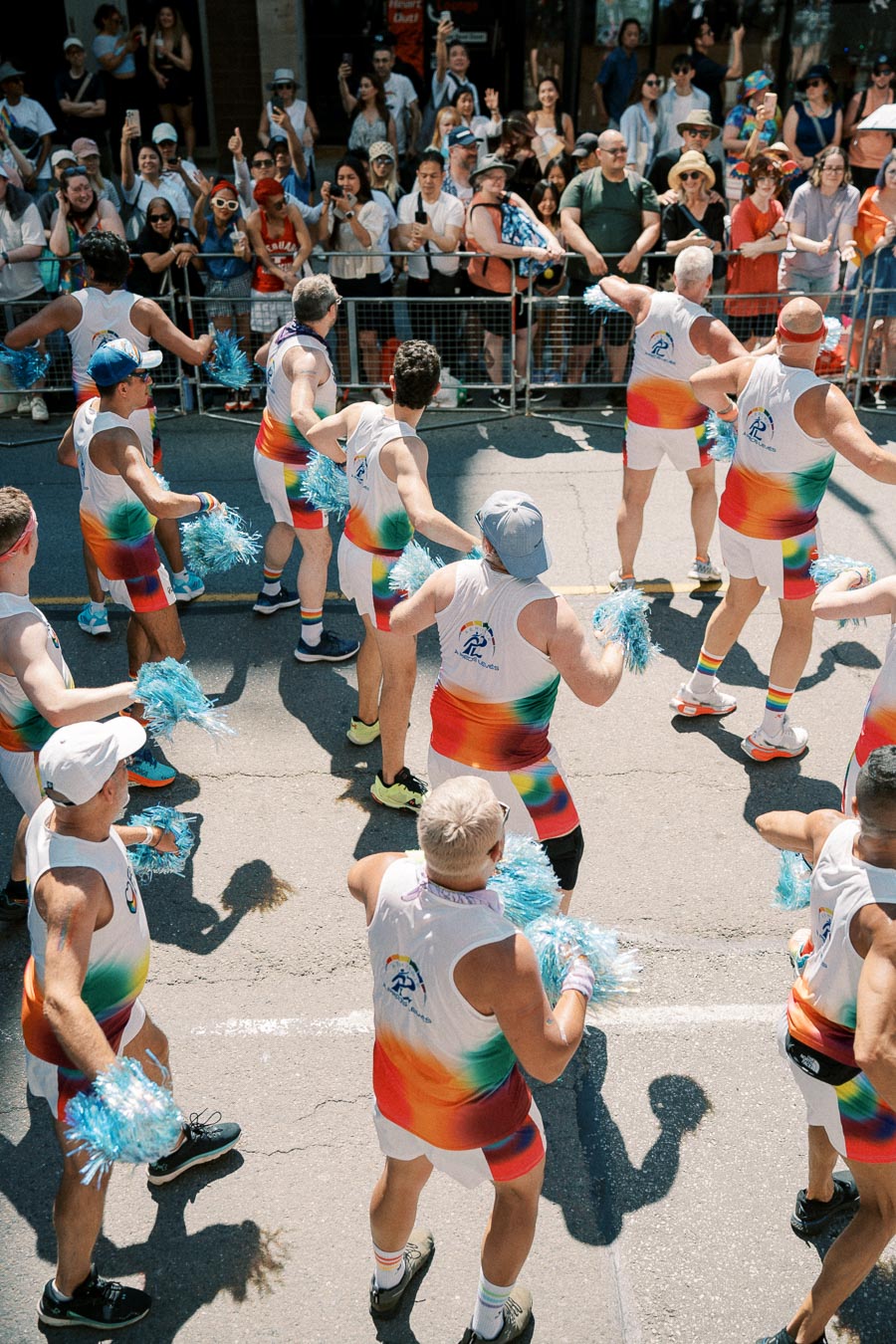 Participants in vibrant rainbow outfits perform with pom-poms during a lively parade, cheered on by an enthusiastic crowd.