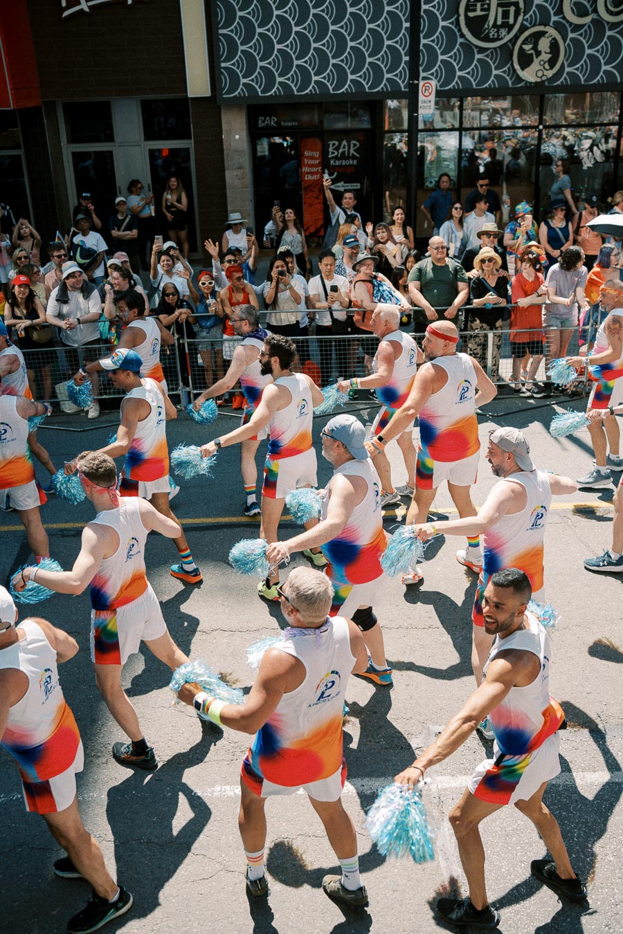 Participants in vibrant rainbow outfits perform with pom-poms during a lively parade, cheered on by an enthusiastic crowd.