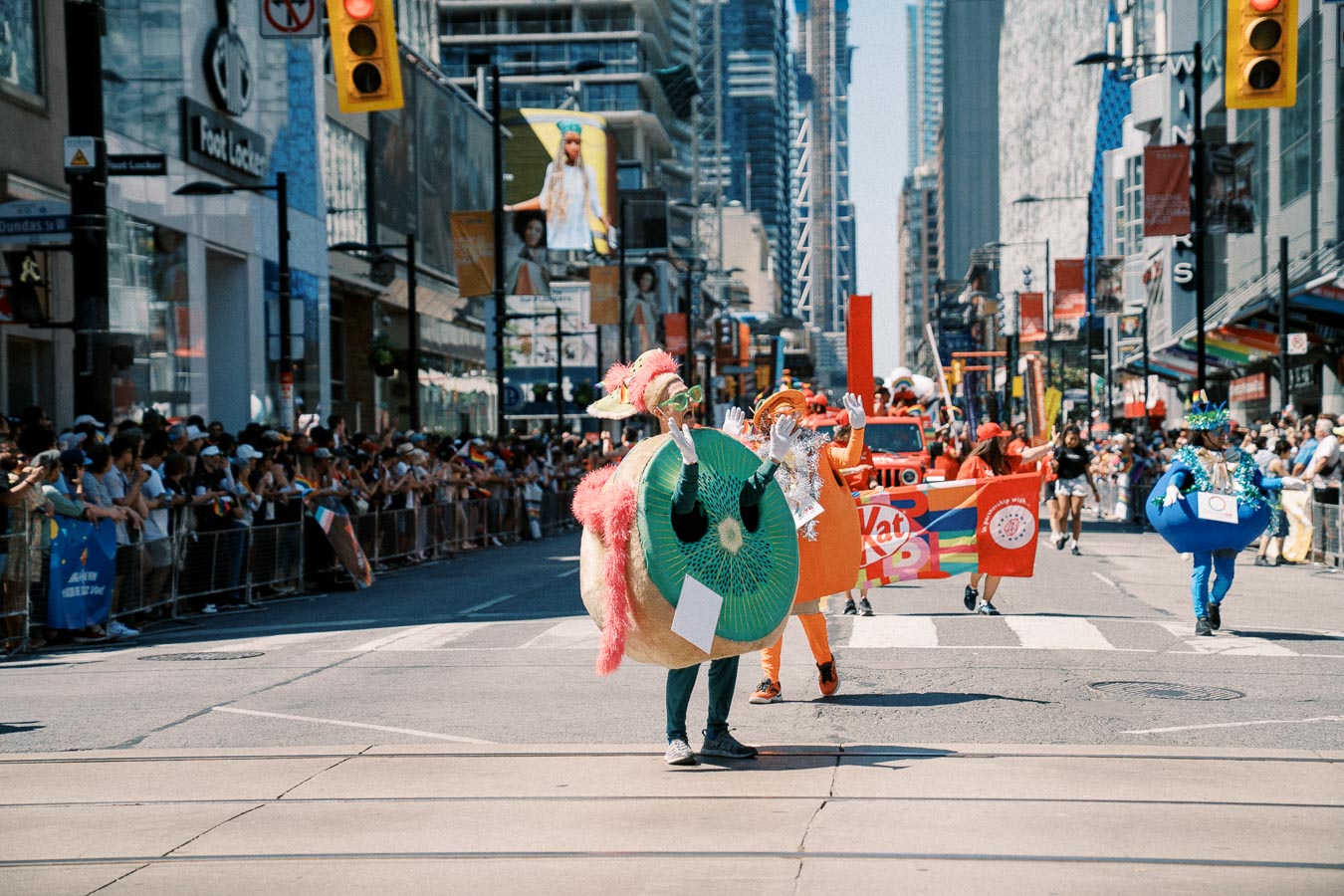 Participants in vibrant costumes, including a kiwi fruit, march in a lively city parade with spectators lining the street.