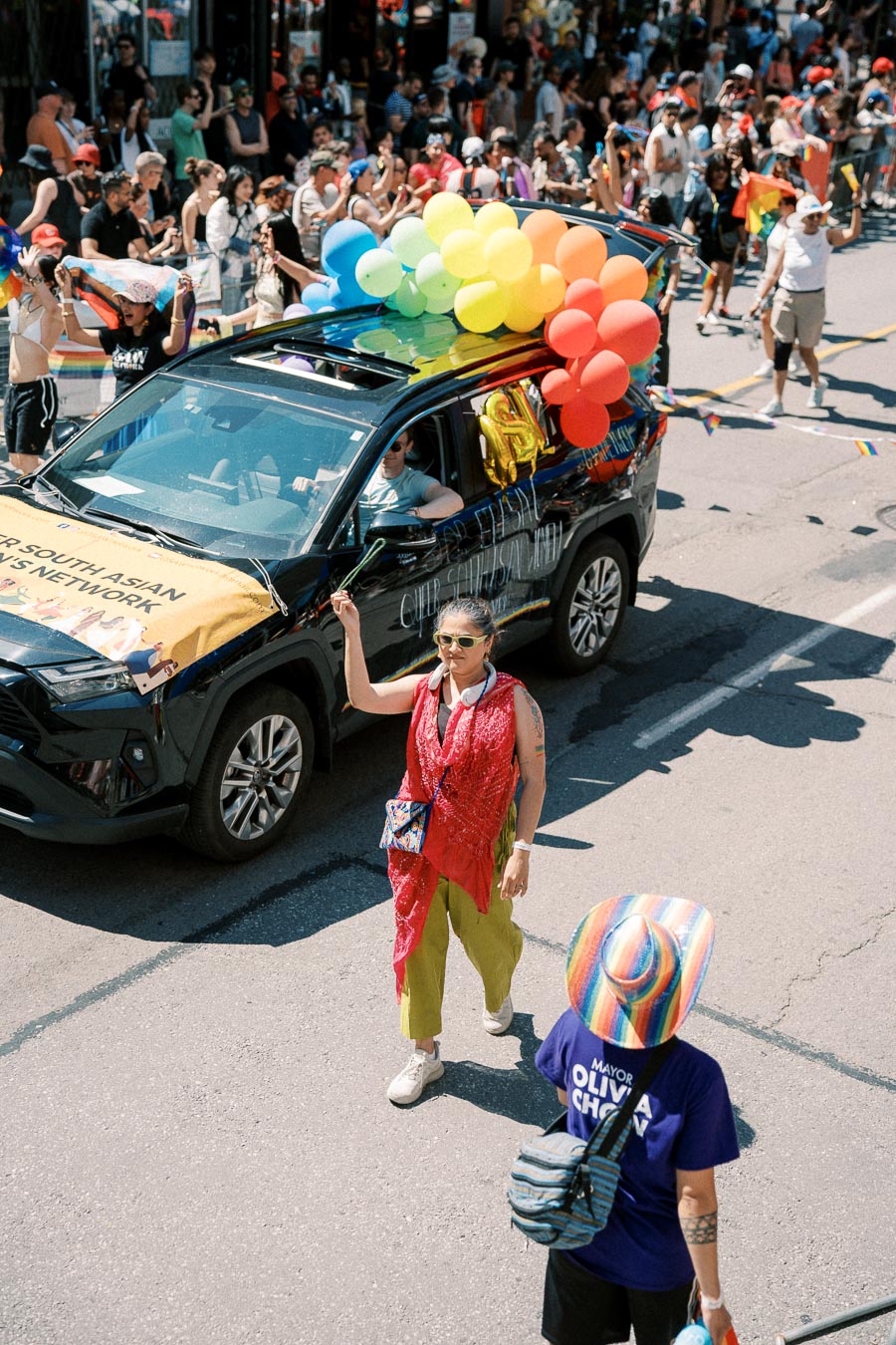 A vibrant parade scene featuring a black car adorned with rainbow balloons and a banner, surrounded by a lively crowd. Participants are celebrating, with one person in a colorful outfit walking alongside the car.