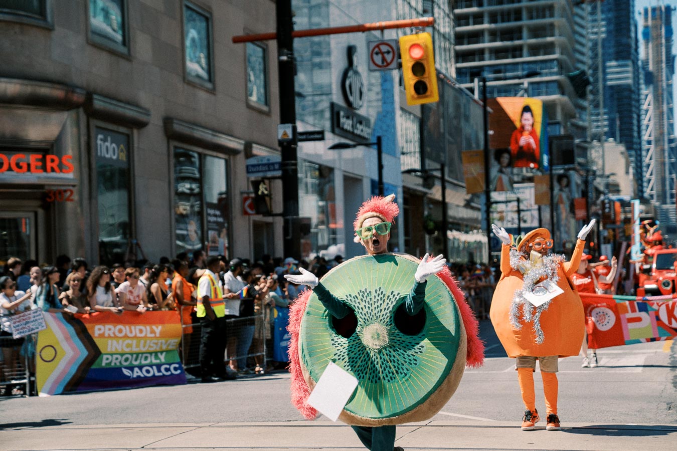 Participants in fruit costumes, including a kiwi and an orange, march in a lively city parade, celebrating diversity and inclusion.