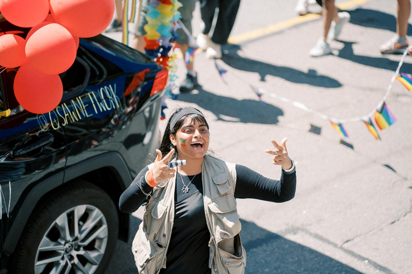 A person participates in a parade, smiling and gesturing with their hands. They have rainbow face paint and are wearing a beige vest over a black shirt. Red balloons are attached to a nearby vehicle, and small rainbow flags are visible in the background.