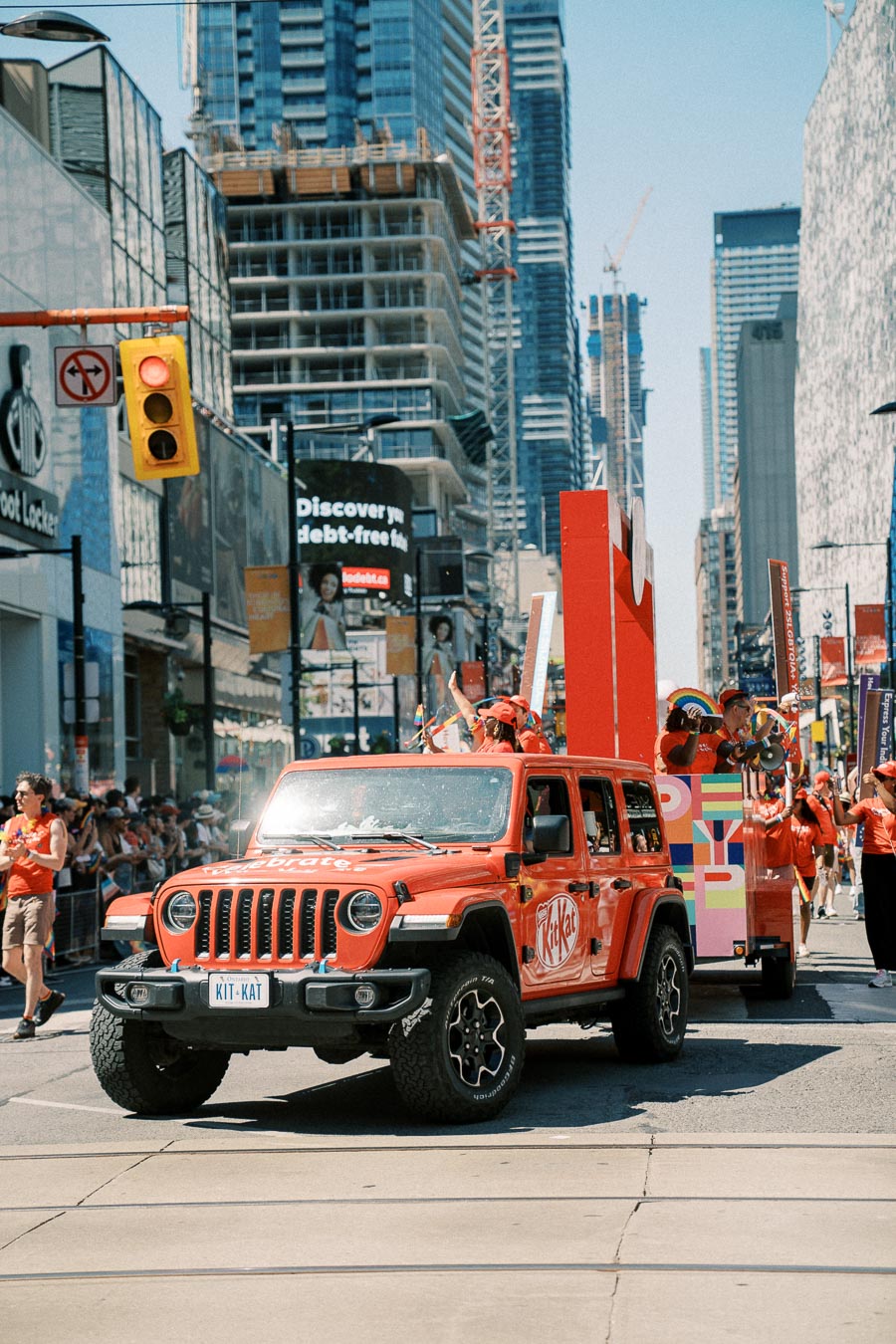 A red Jeep Wrangler with Kit Kat branding participates in a city parade, surrounded by people in red outfits and urban skyscrapers.