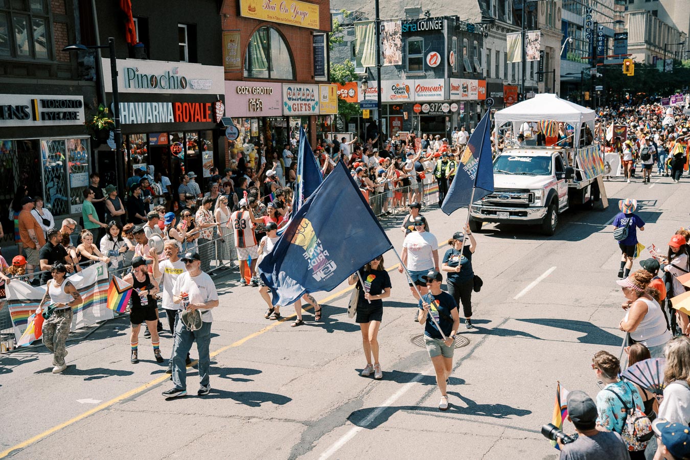 A lively parade scene featuring participants carrying flags and a decorated truck, with spectators lining the street.