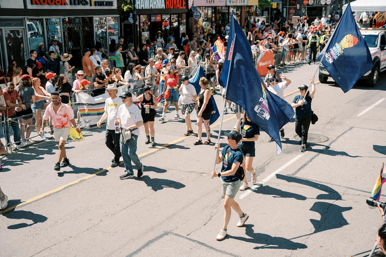 Participants march in a vibrant street parade, carrying flags and celebrating diversity, while a crowd watches from the sidewalk.