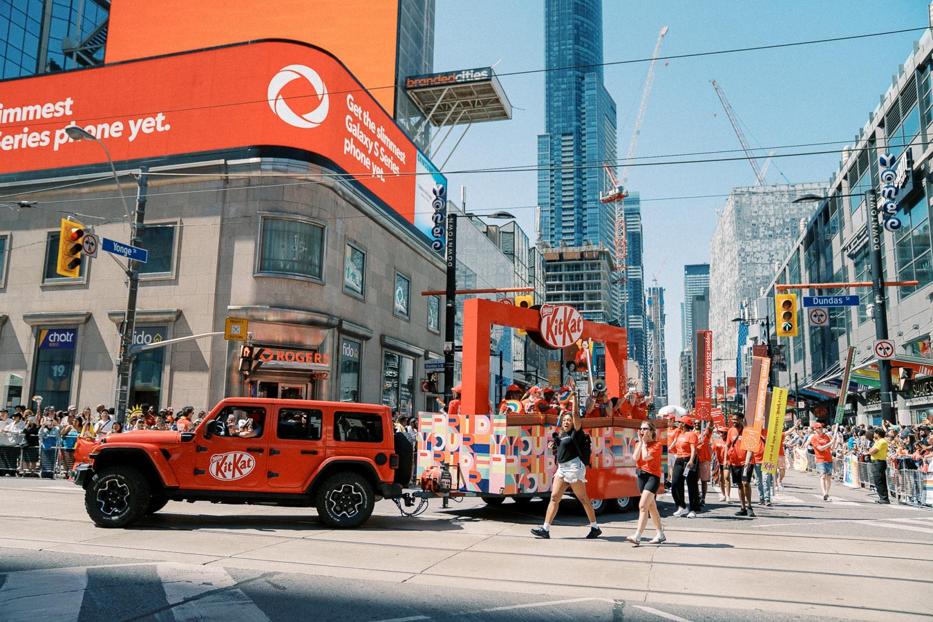 A vibrant parade scene at the intersection of Yonge and Dundas in Toronto, featuring a red Jeep with a KitKat logo towing a colorful float. The street is bustling with people celebrating, and the background is filled with tall buildings and large digital billboards.