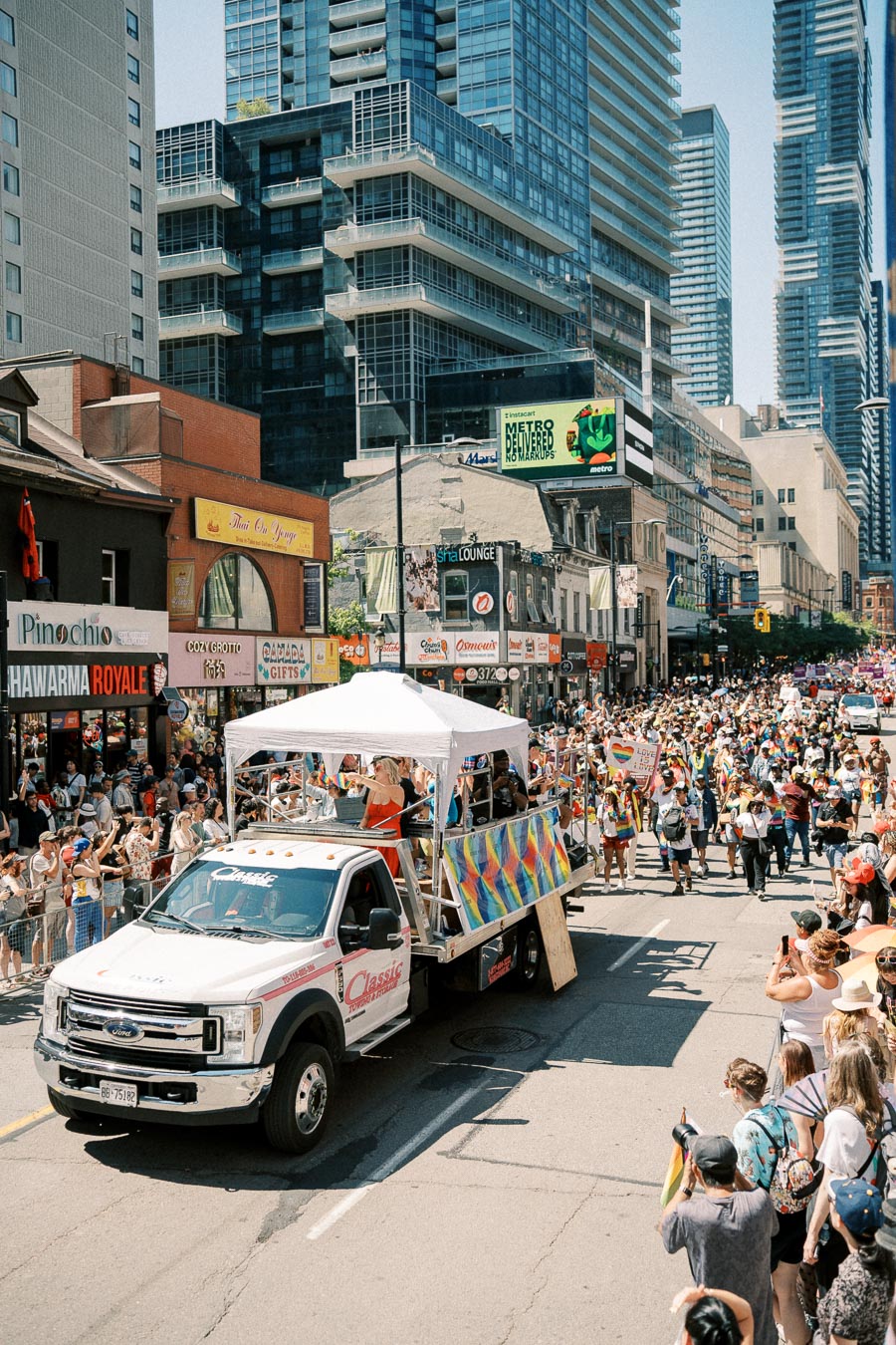 A vibrant parade takes place on a city street, featuring a decorated truck and a large crowd of participants and spectators. Tall modern buildings line the street, adding an urban backdrop to the festive scene.
