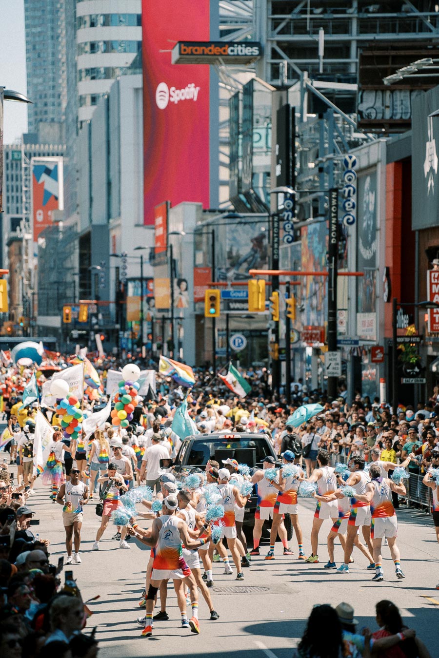 A lively parade takes place on a bustling city street, featuring participants in colorful attire, waving flags and carrying balloons, with a large crowd of spectators lining the sidewalks. The urban backdrop includes tall buildings and prominent advertisements.