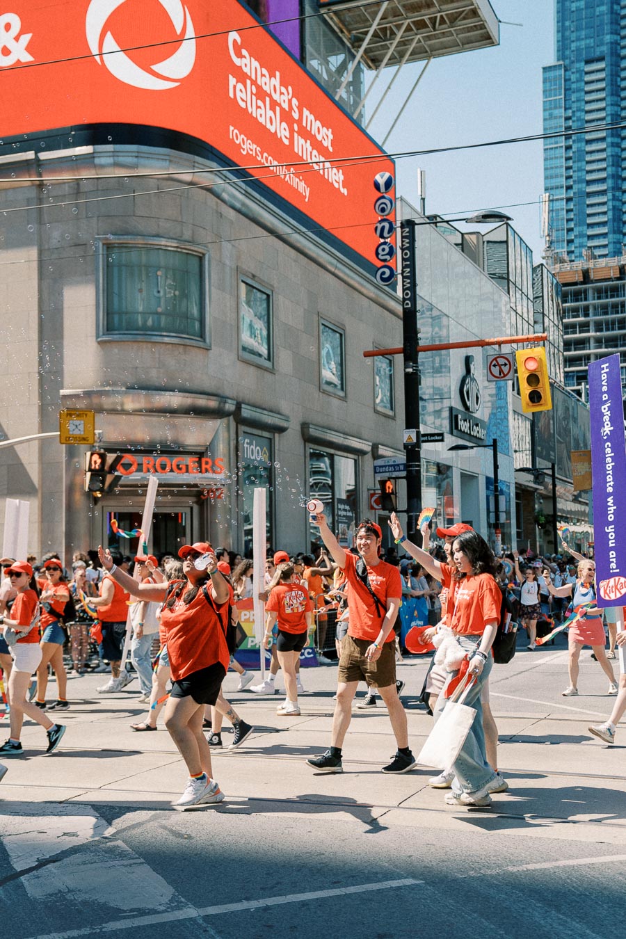 Parade participants in red shirts walk past a Rogers store and a large digital billboard in a bustling city street.