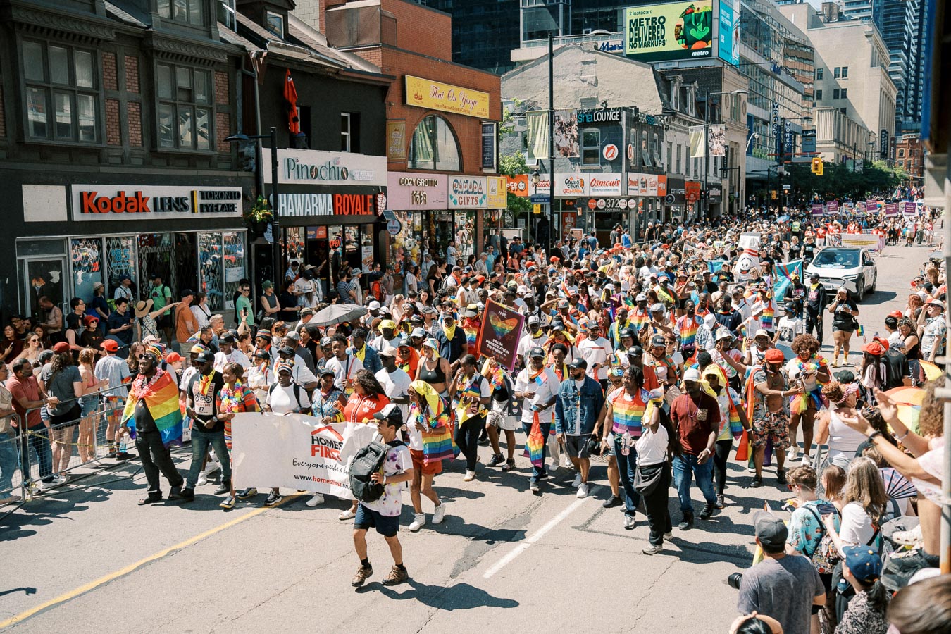 A vibrant pride parade takes place on a bustling city street, with participants waving rainbow flags and carrying banners. Spectators line the sidewalks, enjoying the festive atmosphere.