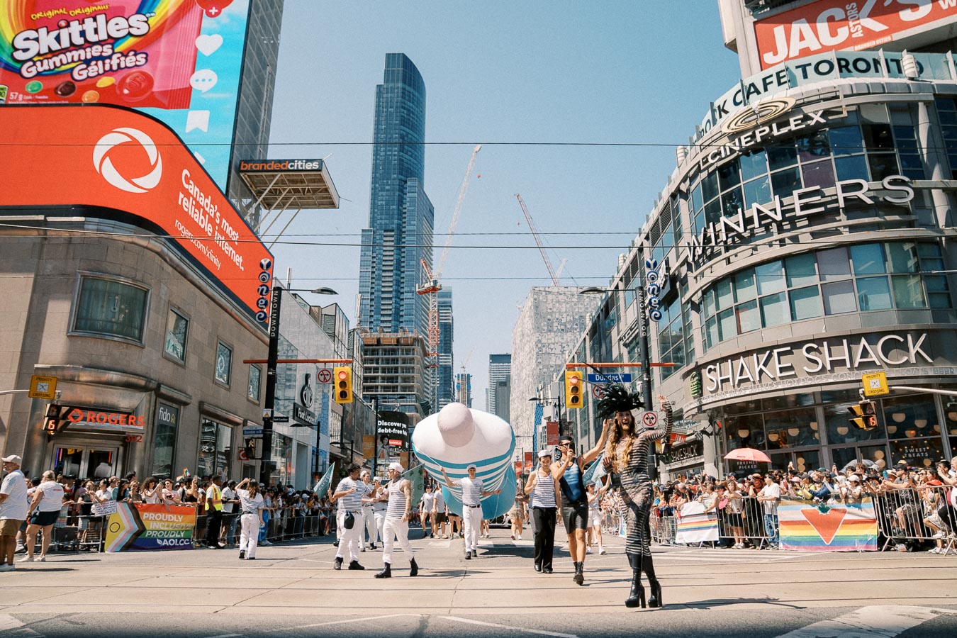 A lively parade in Toronto with participants in costumes and a large inflatable object, set against the backdrop of skyscrapers and bustling storefronts like Shake Shack and Winners.