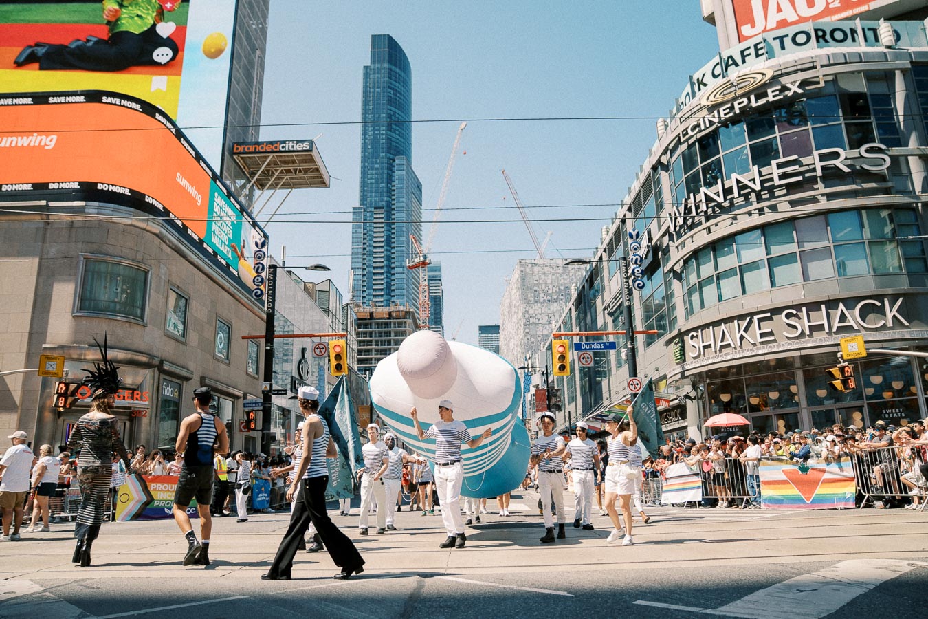 A vibrant parade in a bustling city features participants in striped outfits carrying a large inflatable object. The scene is set against a backdrop of tall buildings, including a Shake Shack and Cineplex, with crowds and rainbow flags lining the streets.