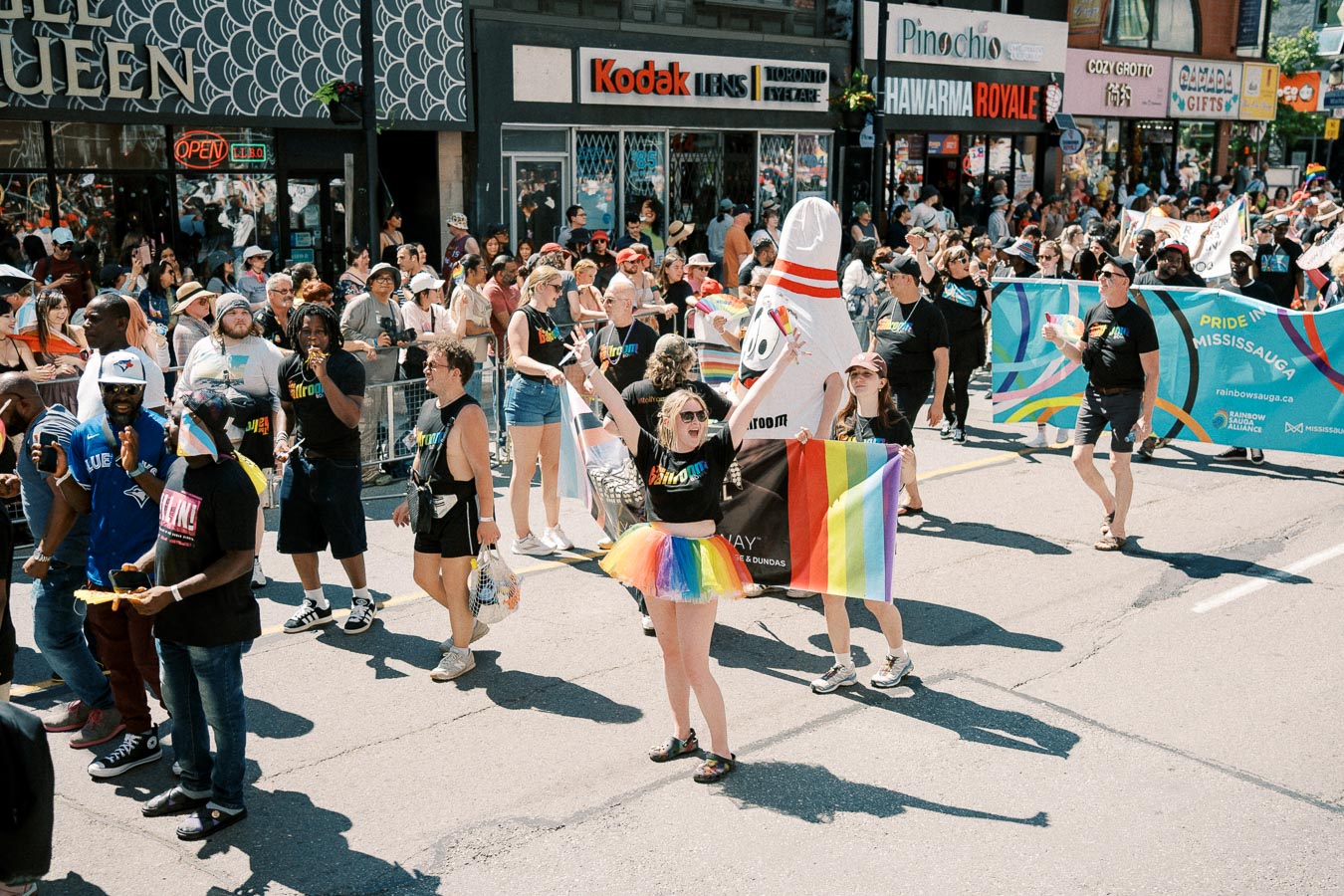 Participants celebrate at a pride parade, holding rainbow flags and a banner for 'Pride in Mississauga'.