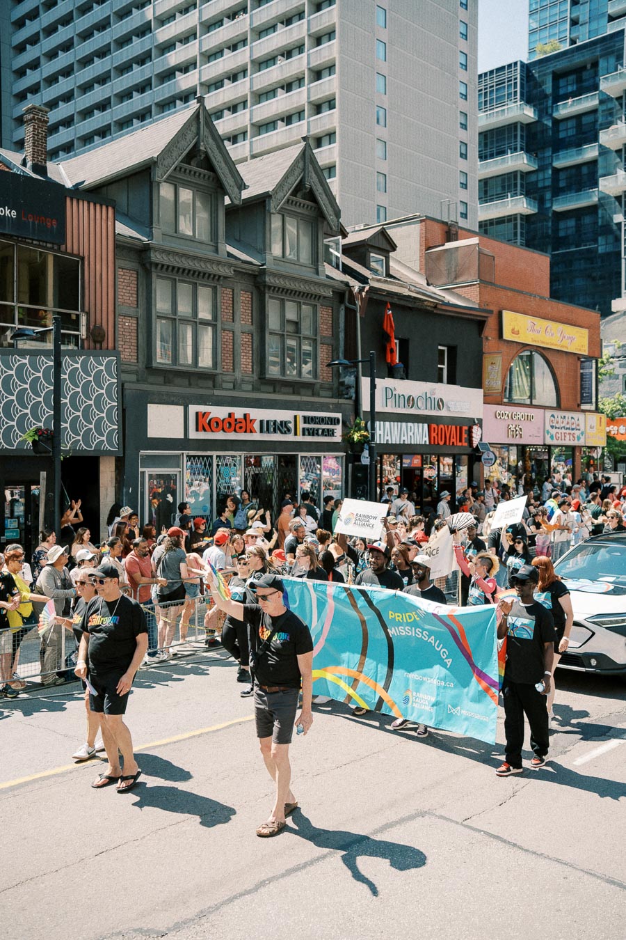 Participants in a Pride parade march down a city street, holding a 'Pride Mississauga' banner, surrounded by onlookers and city buildings.